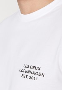 Bílý bavlněný tričko se černým nápisem "LES DEUX COPENHAGEN EST. 2011" na levé straně. Krátké rukávy a kulatý výstřih.