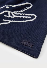 Esarfa tricotată de culoare bleumarin, cu un grafic alb de crocodil și un mic logo brodat în colț. Textură moale, formă dreptunghiulară.