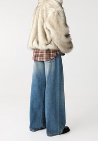 Veste en fausse fourrure crème, superposée à une chemise à carreaux marron, associée à un jean bleu clair à jambes larges. Texture lisse et silhouette décontractée.