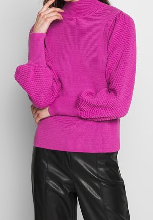 Femme portant un pull tricoté texturé rose vif avec des manches ballon et un pantalon en cuir noir, posant avec la main près du visage.
