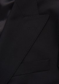 Zwarte blazer met een notch-kraag, textuurstof en een enkele borstzak. Subtiele stiksels langs de randen.