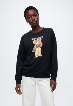 Kvinde iført sort sweatshirt med teddybjørn-grafik, der holder en telefon og en indkøbspose, hvide bukser, mod en ensfarvet hvid baggrund.