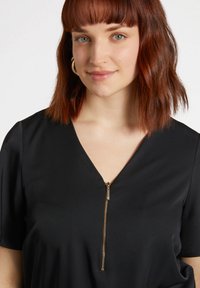 Top negro con escote en V y mangas cortas, con una cremallera frontal en tono dorado. Tejido suave y liso con un ajuste relajado.