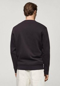 Sudadera negra con puños y dobladillo acanalados, diseño sencillo, cuello redondo, confeccionada con tela suave, combinada con pantalones blancos. Vista trasera.