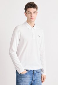 Hvit langermet polo skjorte med krage, med en liten svart logo på venstre bryst og iført lyseblå jeans.