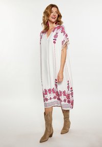 Weißes Kleid mit lebhaftem rosa floralem Stickerei, mit kurzen Ärmeln und einem tiefen V-Ausschnitt. Verziert mit Quastenakzenten am Saum.