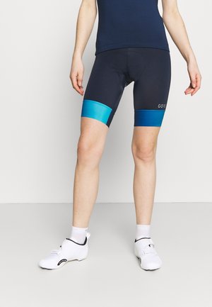 Person trägt schwarze Fahrradhosen mit blauen Beinbändern, weiße Socken und weiße Fahrradschuhe und steht vor neutralem Hintergrund.