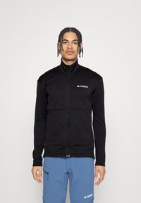 Giovane con capelli intrecciati che indossa una giacca nera Terrex con zip e pantaloni blu Terrex su uno sfondo bianco semplice.