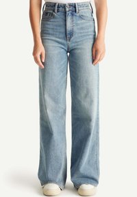 Pantalones vaqueros de tiro alto y pierna ancha en denim azul claro. Presentan un ligero desgastado y un diseño clásico de cinco bolsillos. Combinados con zapatillas blancas.