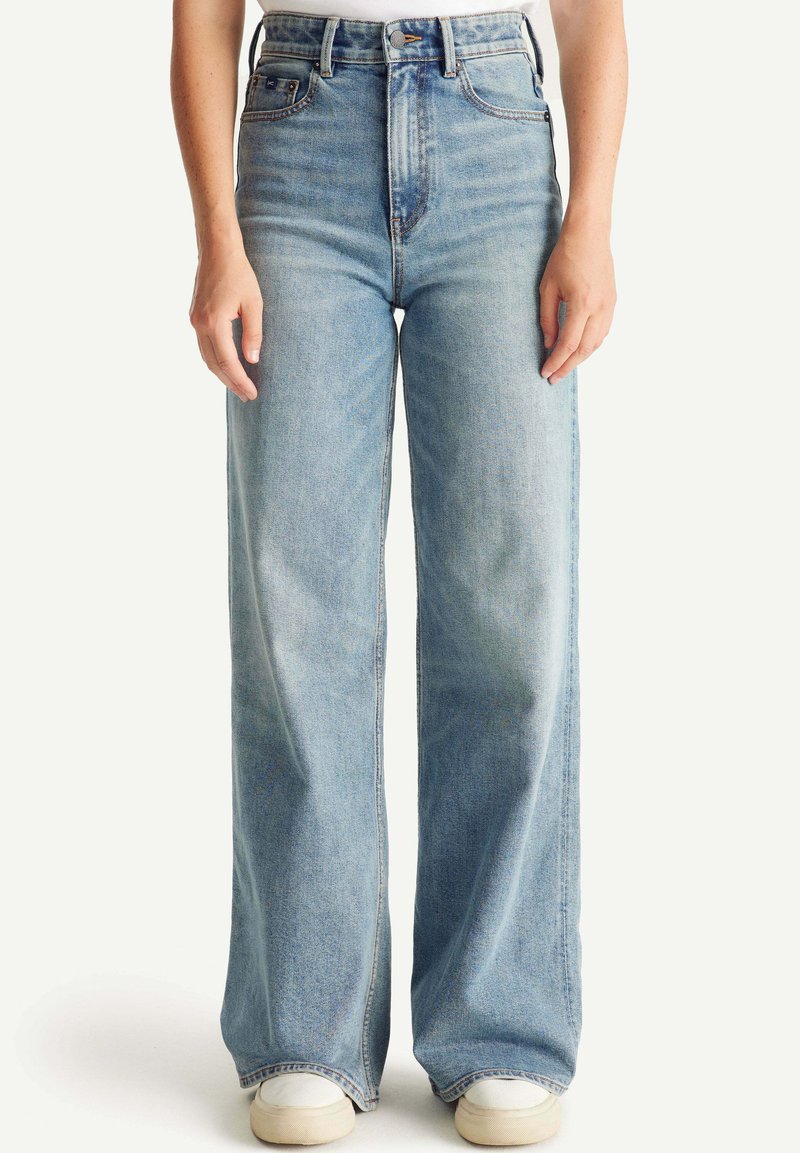 Pantalones vaqueros de tiro alto y pierna ancha en denim azul claro. Presentan un ligero desgastado y un diseño clásico de cinco bolsillos. Combinados con zapatillas blancas.