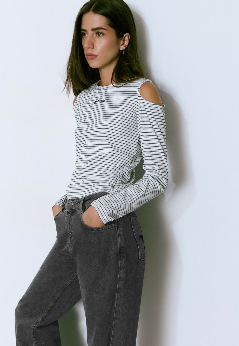 Young woman with long brown hair wears striped cold-shoulder top and dark high-waisted jeans, standing with hands in pockets against white wall.