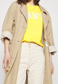 Gabardina beige con las mangas enrolladas sobre una camiseta amarilla brillante con letras blancas, combinada con jeans de color claro.