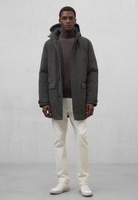 Ciemnozielona parka z kapturem i kieszeniami, noszona na brązowym swetrze z golfem. Do tego białe spodnie i białe tenisówki.