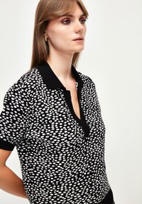 Černo-bíle vzorovaná polokošile s krátkým rukávem, límečkem a knoflíkovou légou, která má texturovaný pletený design.