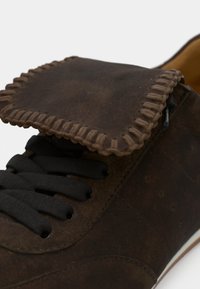 Καφέ δερμάτινο παπούτσι με χοντρές μαύρες κορδόνια και λεπτομερή ραφή whipstitch στη διπλωμένη γλώσσα σε λευκό φόντο.