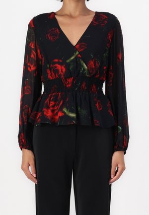 Frau trägt eine schwarze Bluse mit roten Rosenmustern, langen transparenten Ärmeln, einem V-Ausschnitt und einem gerafften Taillenbund, kombiniert mit schwarzen Hosen.
