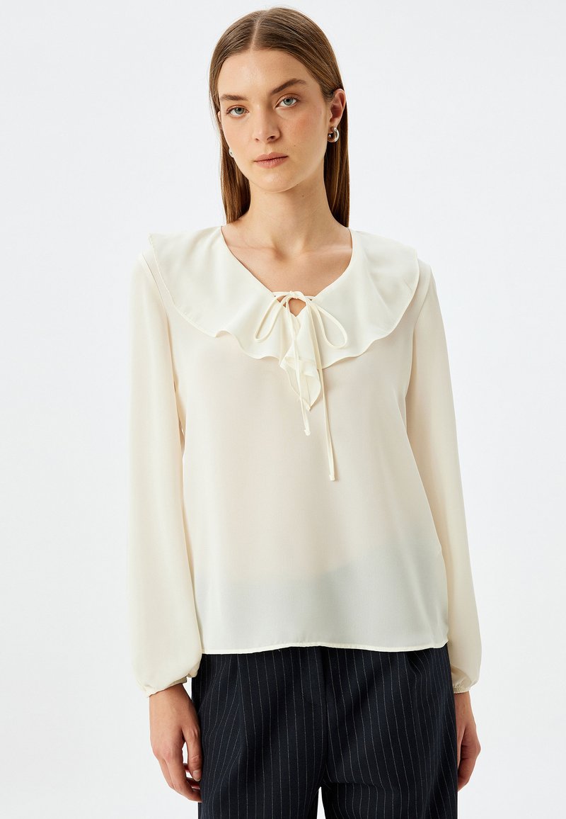Creme bluse med opsvulmede lange ærmer, en flæsset krave og en sløjfedetalje ved halsen. Let, glat stof med en afslappet pasform.