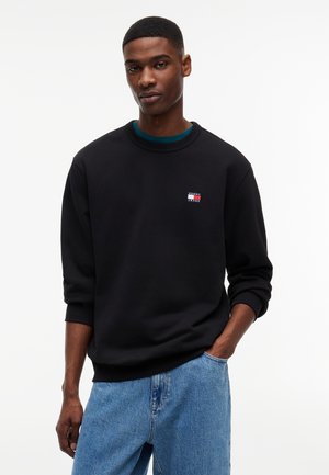 Mand iført sort Tommy Jeans sweatshirt og blå jeans, stående med den ene hånd i lommen mod en hvid baggrund.