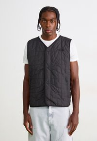 Gilet matelassé noir avec un col en V, fermeture par pression à l'avant et poches zippées. Porté sur une chemise blanche, arborant un logo discret sur la poitrine.