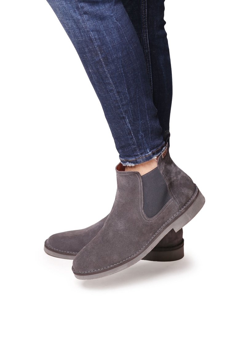 Personne portant un jean slim bleu foncé et des bottines Chelsea gris en suède avec des panneaux latéraux élastiques sur fond blanc.