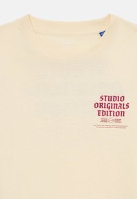 Svetle béžové tričko s kulatým výstřihem. Tisknutý text v tmavě červené barvě říká "STUDIO ORIGINALS EDITION" a pod ním je další menší text.