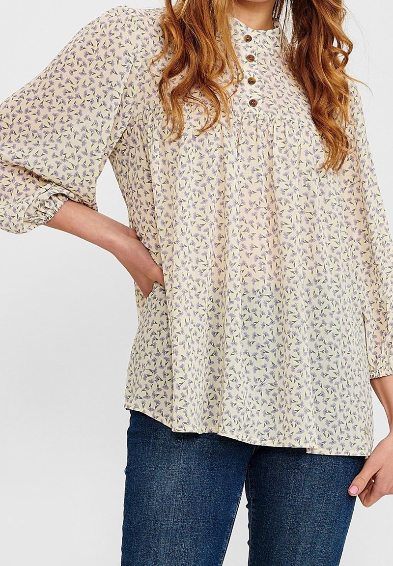 Hellästi kuvioitu pusero pehmeästä kankaasta, jossa on korkea kaulus, napitus ja kerätyt hihat. Värit sisältävät vaaleankeltaista ja violetteja kukkia.