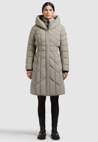 Woman wearing a knee-length, padded, light gray coat with a high collar, paired with black leggings and black ankle boots.