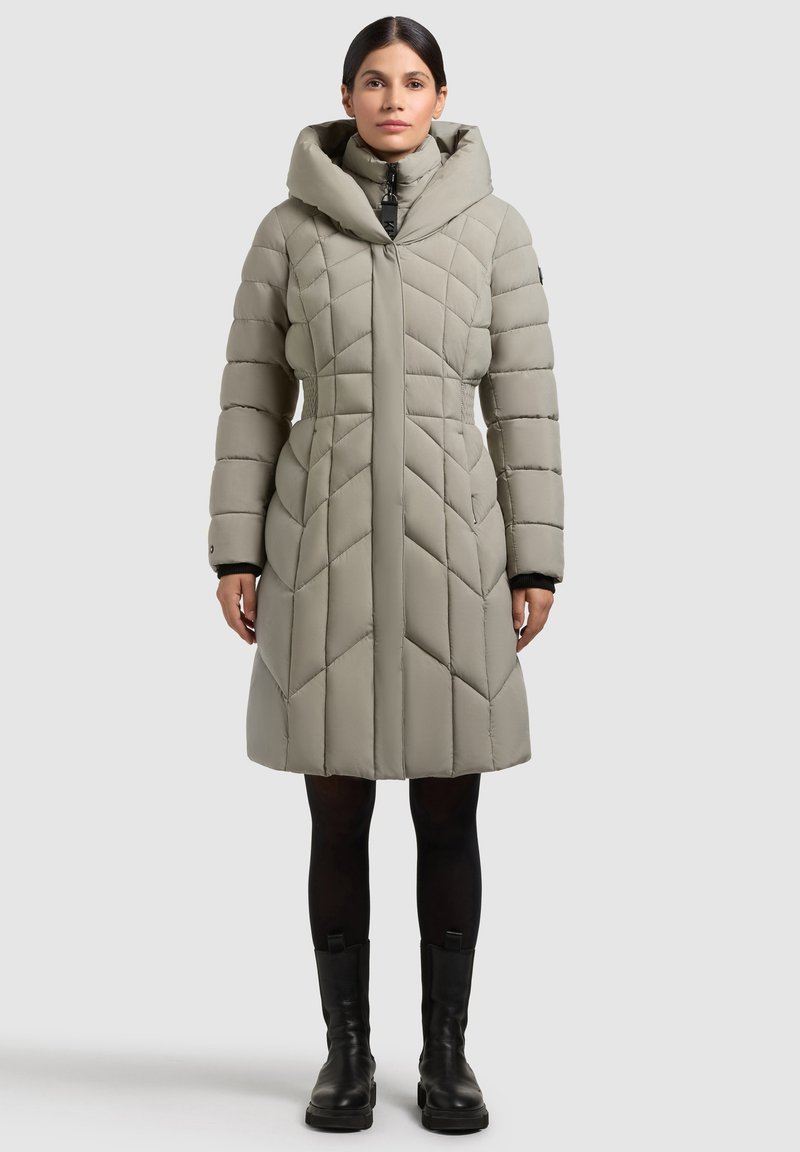 Woman wearing a knee-length, padded, light gray coat with a high collar, paired with black leggings and black ankle boots.