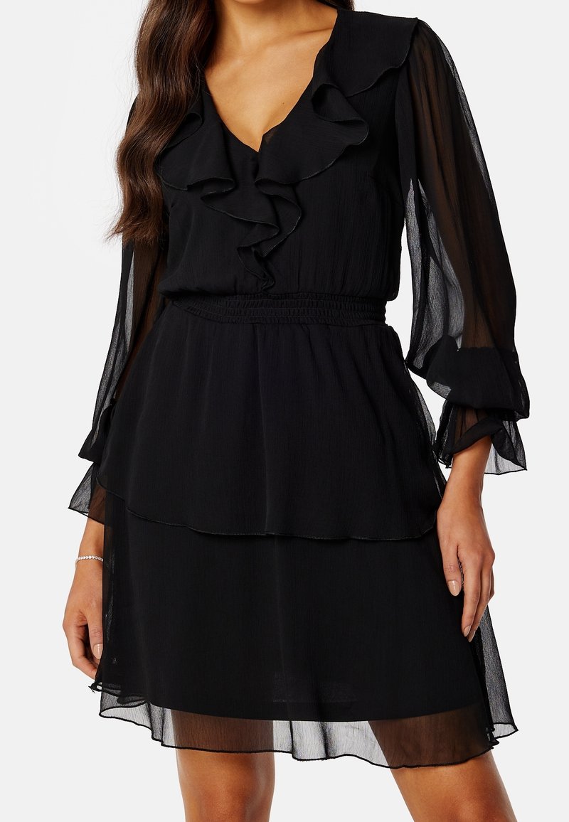 Femme portant une robe noire à manches longues avec volants, col en V, manches transparentes et jupe à étages, debout avec les bras détendus.