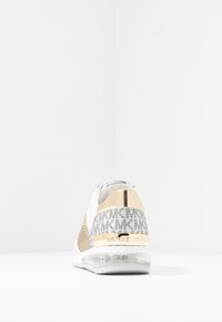 Zapatilla deportiva blanca con acentos dorados, con una parte superior de malla, panel de logo plateado texturizado y suela transparente. La vista trasera muestra el detalle del logo.