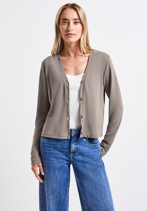 Khakifarbenes Strickjacke mit V-Ausschnitt, Knopfverschluss und langen Ärmeln, getragen über einem weißen Oberteil und kombiniert mit hochgeschnittenen blauen Jeans.