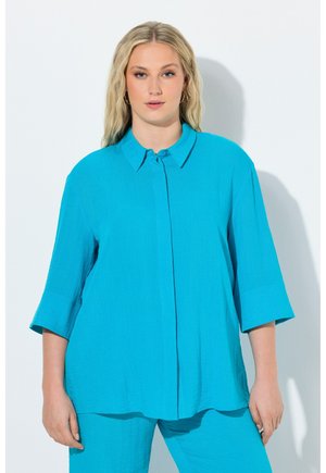 Blonde vrouw die een losse turquoise blouse met knopen en driekwart mouwen draagt, samen met bijpassende broek, staand tegen een witte achtergrond.