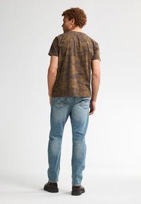 Bruin patroon t-shirt met korte mouwen, gecombineerd met lichtblauwe jeans. Het model staat met de rug naar de camera, wat een ontspannen pasvorm en kraagontwerp laat zien.