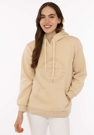 Junge Frau mit langen braunen Haaren, die einen beigen Hoodie mit Text und Herz-Design trägt, steht mit einer Hand in der Tasche vor einem schlichten Hintergrund.