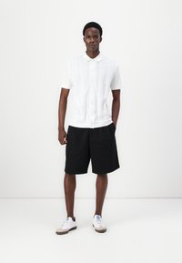 Balta īsām piedurknēm pogājama krekla ar teksturētu rakstu, kas saskaņots ar melniem šortiem un baltiem sporta apaviem, ar ziliem akcentiem un gaišām zālēm.