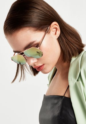 Gafas de sol con montura de metal rosa y lentes espejo verdes. El modelo tiene el cabello castaño y lleva una chaqueta verde clara sobre una camiseta negra.
