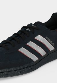 Σκούρο μπλε αθλητικό παπούτσι Adidas με μαύρα κορδόνια, τρεις αντανακλαστικές ασημένιες ρίγες με κόκκινη περίμετρο και λογότυπο "Messi" στο πλάι.