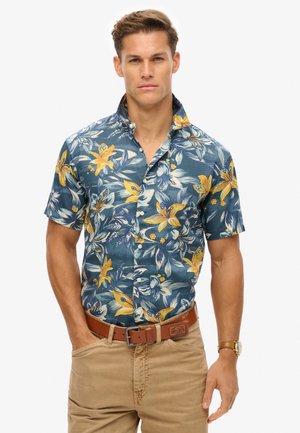 Camisa de manga corta azul marino con patrón floral amarillo, de diseño abotonado y con cuello. Combinada con pantalones caqui claros y un cinturón marrón.