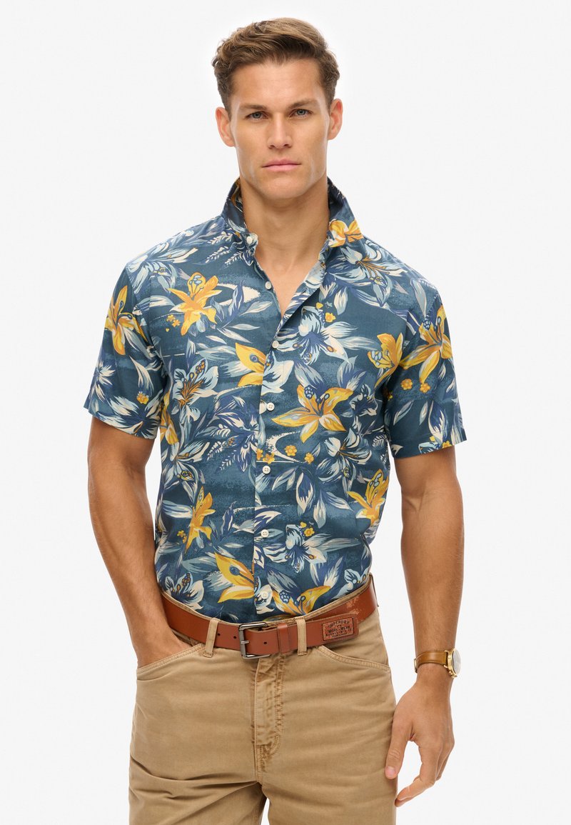 Camicetta azzurra scura a maniche corte con motivo floreale giallo, design con bottoni e colletto. Abbinata a pantaloni beige chiaro e una cintura marrone.