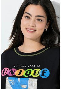 Camiseta negra con cuello redondo y costuras en verde neón; presenta un texto colorido "TODO LO QUE NECESITAS ES ÚNICO" con diseño gráfico.