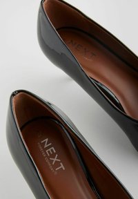 Musta lakeri ballerinat, joissa on pyöreä nenä, sileä pinta ja ruskea sisus, jossa on sisällä 'NEXT FOREVER COMFORT' -brändäys.