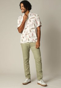 Kortebloemshirt in het wit met roze en groene bladerenpatronen, gecombineerd met lichtgroene broek en witte sneakers met bruine zolen.
