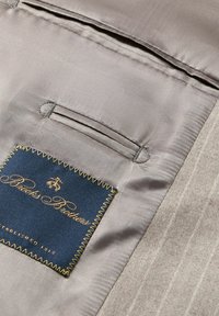 Interno in tessuto grigio con una sola tasca e un'etichetta blu con scritte dorate che dice "Brooks Brothers", evidenziata da cuciture gialle.