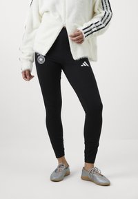 Leggings pretas com logótipos e riscas brancas. Uma jaqueta felpuda branca com mangas com riscas pretas. Sapatos prateados com atacadores e solas de borracha.