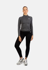 Šedá atletická bunda na zips s dlhými rukávmi, kombinovaná s čiernymi legínami a bielymi teniskami. Hladká textúra, priliehavý strih a logo akcenty.