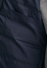 Tela sintética color azul marino, brillante, con un acabado elegante. Cuenta con un bolsillo de parche y una tela gris texturizada adyacente.