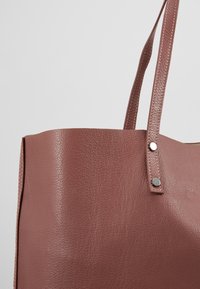 Ruusuin värinen nahkainen olkalaukku, jossa on sileä pinta, pystysuuntaiset saumat ja kaksi kantohihnaa. Sivulla on napsautettava suljin.