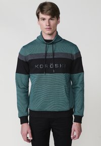 Sudadera verde con cuello alto, con rayas horizontales en negro y gris, un bolsillo frontal y el texto "KORÖSHI" en el pecho.