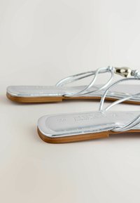 Sandales plates argentées avec fines lanières et un petit détail de perle métallique, dotées d'une semelle marron et d'un design à bout carré.