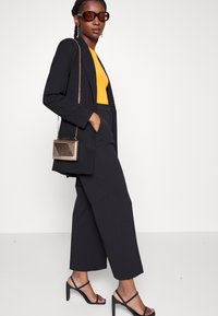 Blazer noir ajusté sur un haut jaune, associé à un pantalon noir à jambes larges. Les accessoires incluent des lunettes de soleil brunes oversized et une pochette à motifs.